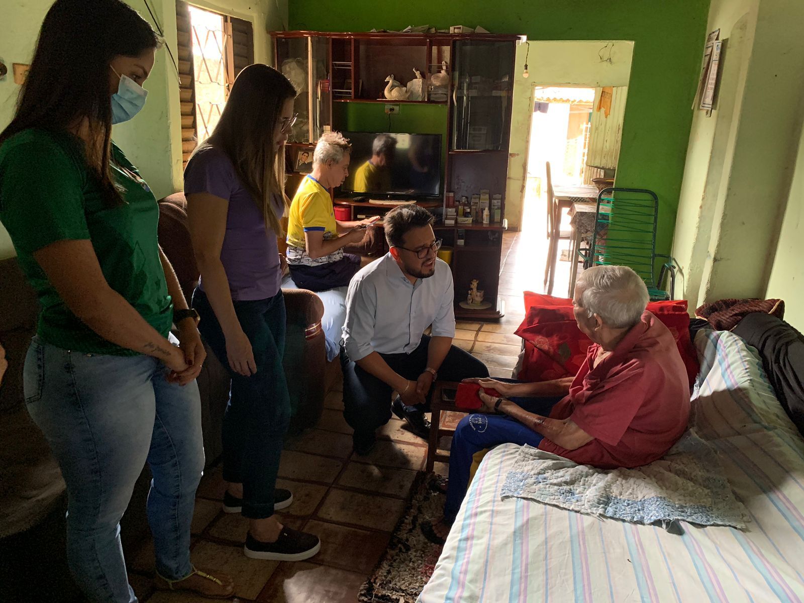 Foto na horizontal feita na sala de uma casa com paredes verdes. No centro, o secretário de saúde Gustavo Queiroz está agachado de frente para o senhor Jovelino, um homem idoso sentado de maneira curvada em um sofá coberto com um lençol. à esquerda de Gustavo Queiroz, está a secretária de saúde Rouane Azevedo, em pé. Ao lado dela, uma funcionária de prefeitura, uniformizada e de máscara.